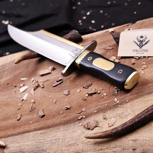 D2 Iron Mistress bowie knife with brass fittings and black G10 handle angled on wooden board