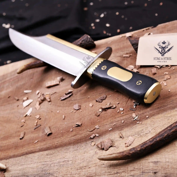 D2 Iron Mistress bowie knife with brass fittings and black G10 handle angled on wooden board