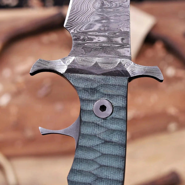 Close-up macro shot of Damascus steel bowie knife showing double finger guard, ricasso pattern, and textured green handle