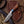 A fixed-blade knife with a patterned steel blade, a brown wood handle with a blue accent, and a leather lanyard, displayed diagonally on its brown embossed leather sheath over a rustic wooden surface with straw accents.
