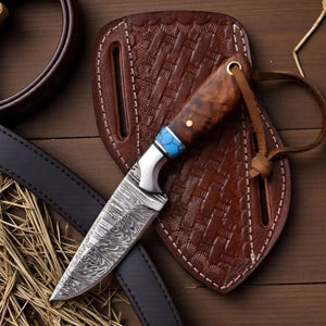 A fixed-blade knife with a patterned steel blade, a brown wood handle with a blue accent, and a leather lanyard, displayed diagonally on its brown embossed leather sheath over a rustic wooden surface with straw accents.