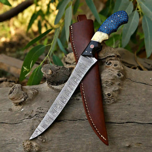 A fixed-blade knife with a patterned steel blade, black handle, and blue inlay, resting diagonally on a brown leather sheath against a rustic wood log with green foliage in the background.