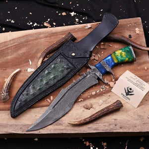 A custom Finger Guard Bowie knife with a San Mai Damascus steel blade, black leather sheath, and a colorful green and blue wood handle, displayed on a rustic wooden board 