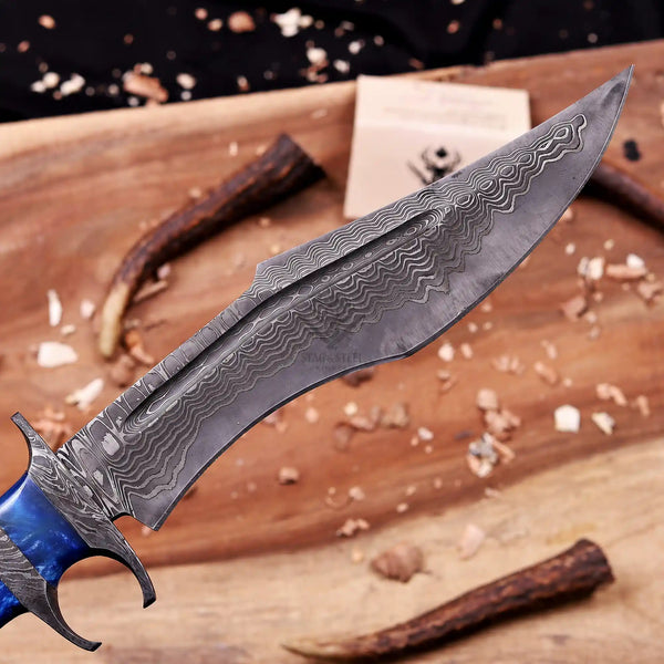 A Close up of San Mai Damascus steel blade knife with a beautiful patterned blade and a blue handle, displayed diagonally on a wooden cutting board with wood shavings and antlers.