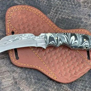 A fixed-blade damascus hawkbill knife with a patterned silver handle and a curved Damascus-style blade, resting on a brown woven leather sheath over a textured gray surface.