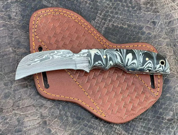 A fixed-blade knife with a patterned steel blade and a black-and-white marbled handle, resting on a brown woven leather sheath over a textured gray surface.