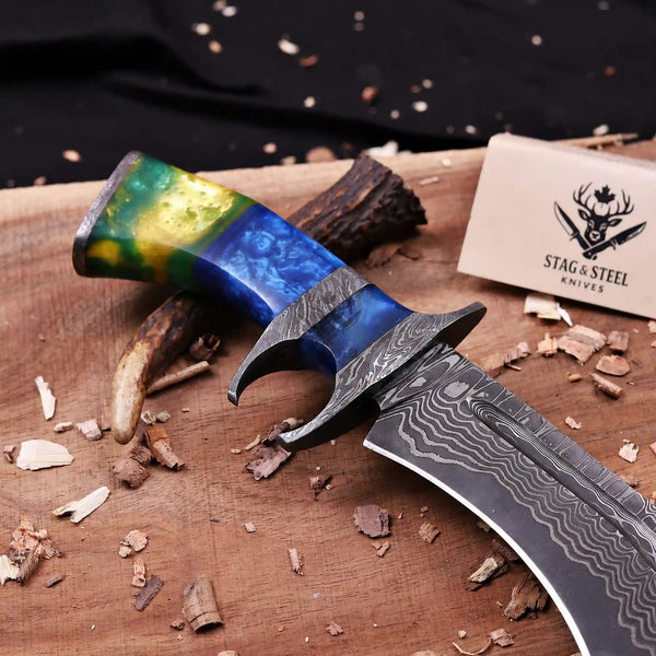 A custom handcrafted knife with a patterned Damascus steel blade and a vibrant marbled blue, green, and yellow handle, displayed on a wooden surface with wood shavings and a Stag & Steel Knives brand tag.