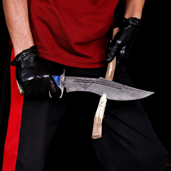 A close-up of a person wearing black gloves and a red shirt, holding a large Finger Guard Bowie knife with a San Mai Damascus steel blade and resin handle cutting the firewood.