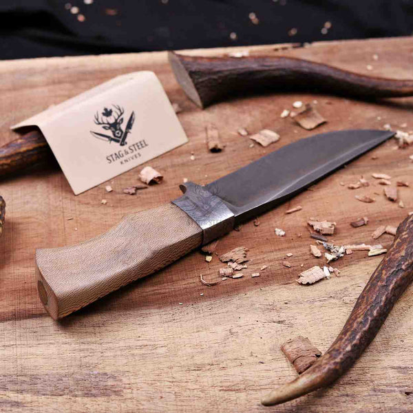 A fixed-blade knife with a textured tan handle and a dark steel blade lies on a wooden surface with wood shavings. A branded card reading STAGE STEEL SWISS is visible, along with antler-like handles or tools in the background.