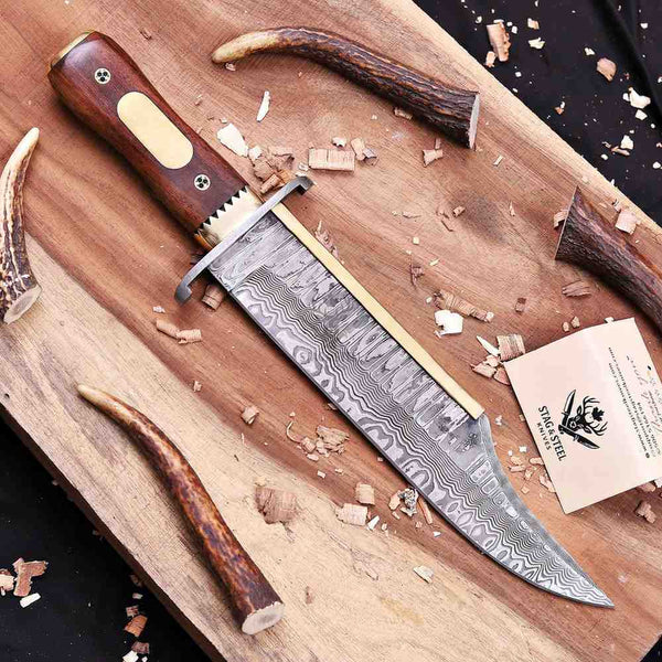 A custom fixed-blade knife with a Damascus steel blade and a reddish-brown wooden handle, displayed diagonally on a wooden cutting board with wood shavings, antlers, and a brand card.