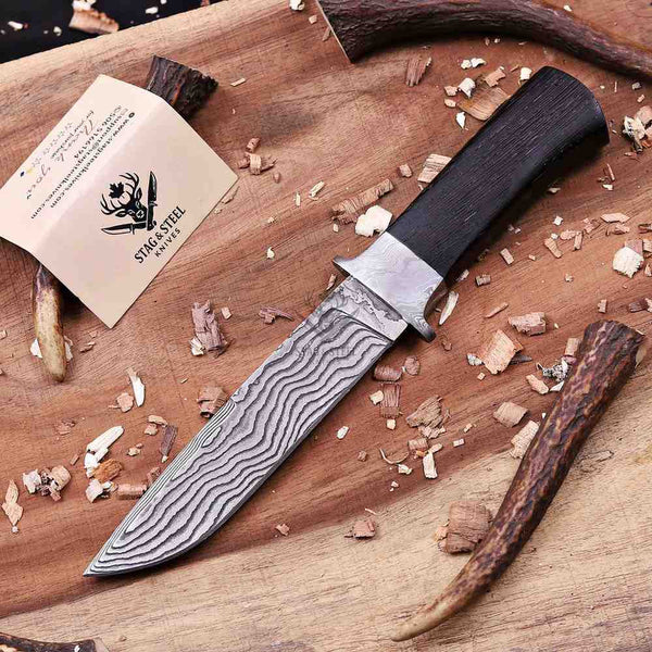 A fixed-blade knife with a patterned Damascus steel blade and a dark wood handle, displayed on a wooden cutting board with wood shavings and antler props. A branded paper tag reading 'STRAIGHTEN' is visible near the top left.