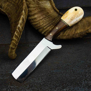 A fixed-blade knife with a polished steel blade and a dual-tone handle made of dark brown wood and cream-colored horn, resting on a dark textured surface with a dried animal horn prop.