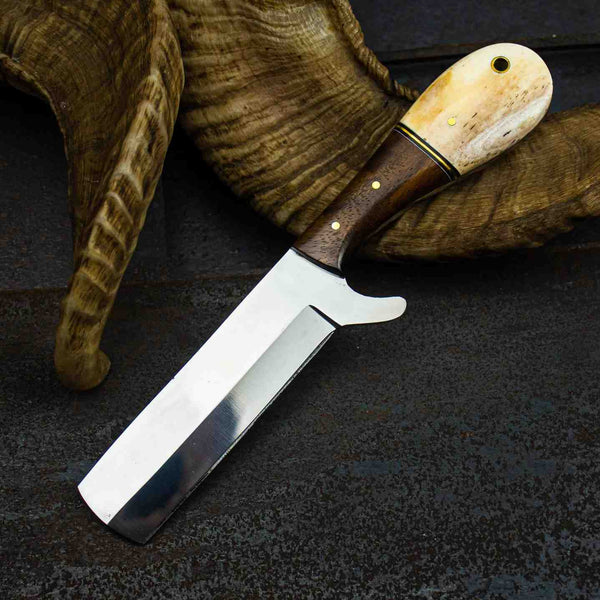 A fixed-blade knife with a polished steel blade and a dual-tone handle made of dark brown wood and cream-colored horn, resting on a dark textured surface with a dried animal horn prop.