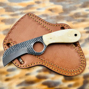 A fixed-blade knife with a textured dark gray blade and a cream-colored handle, resting on a brown leather sheath over a leopard-print fabric background.