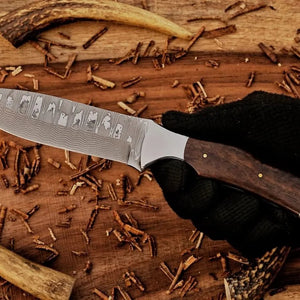 Knife with a wooden handle held by a gloved hand on a wooden surface with dried leaves.