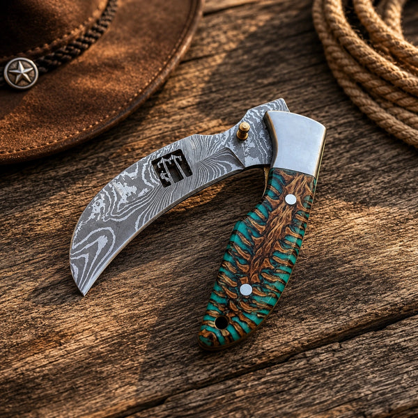 A custom folding knife with a curved Damascus steel blade and a textured handle featuring turquoise and brown wood inlay, displayed on a rustic wooden surface with a cowboy hat and rope in the background.
