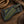A custom handcrafted leather knife sheath with a geometric diamond pattern in green and tan, featuring white contrast stitching and a wrapped knife handle, displayed on a rustic wooden surface.