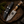 A fixed-blade knife with a patterned steel blade and a carved handle, displayed beside a black leather sheath on a rustic wood slice with pine branches and dark stones.