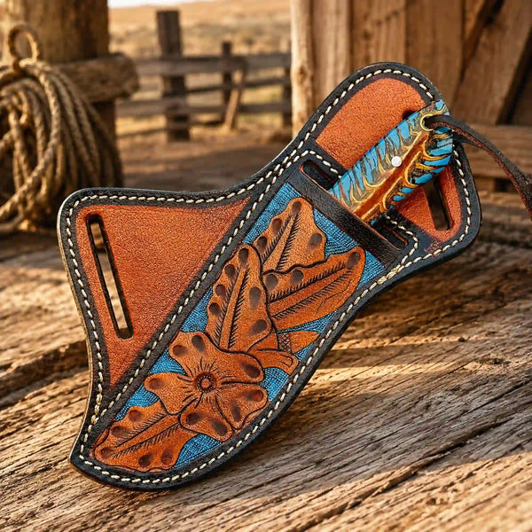 A hand-tooled leather knife sheath with floral engravings, a turquoise and blue feather accent, and white stitching, resting on a rustic wooden surface.