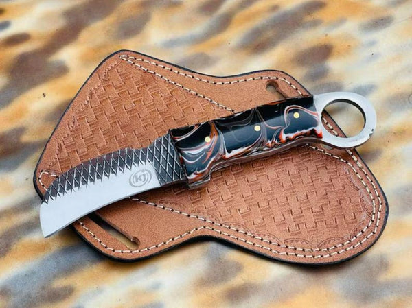 Custom handmade rasp steel hawkbill knife with a black, orange, and white swirled resin handle and ring pommel, resting on a light brown basket-weave leather sheath.