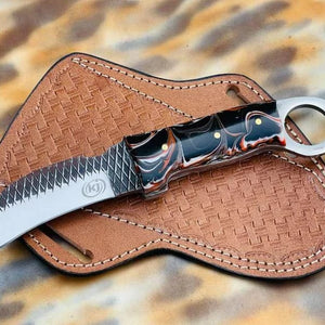 A hawkbill knife with a black and copper marbled handle and a partially serrated blade, resting on a brown woven leather sheath over a leopard-print background.