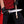 A close-up of a person wearing a red shirt and black pants, holding a large fixed-blade knife with a silver blade and brown wooden handle. The person is also wearing black gloves, and the background is solid black.