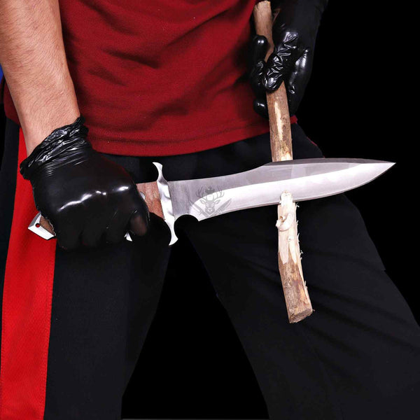 A close-up of a person wearing a red shirt and black pants, holding a large fixed-blade knife with a silver blade and brown wooden handle. The person is also wearing black gloves, and the background is solid black.