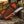 A fixed-blade knife with a polished steel blade and brown wooden handle rests against its matching brown leather sheath on a stone surface, with a rustic outdoor background.