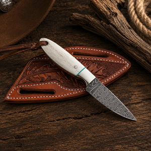 A small fixed-blade knife with a patterned steel blade and white handle rests on a brown leather sheath, placed on a rustic wooden surface with a cowboy hat, rope, and wood props nearby.