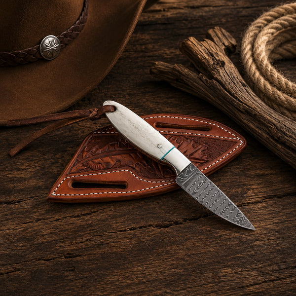 A small fixed-blade knife with a patterned steel blade and white handle rests on a brown leather sheath, placed on a rustic wooden surface with a cowboy hat, rope, and wood props nearby.