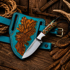 A handcrafted fixed-blade knife with a turquoise leather sheath featuring embossed floral tooling, displayed on a rustic wooden surface with rope and leather gloves as props.