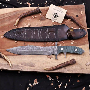 A fixed-blade knife with a patterned steel blade and a textured green handle is displayed on a wooden board alongside its dark brown leather sheath, antlers, wood shavings, and a brand card for 'SikuAte Knife Co.'.