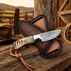 A fixed-blade knife with a patterned steel blade and a marbled bone handle rests on a brown leather sheath, set on a rustic wooden surface with a blurred western background.