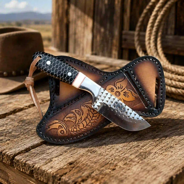 A fixed-blade knife with a textured silver blade and honeycomb-patterned handle, resting inside a brown leather sheath with floral tooling, set on a rustic wooden surface with a lasso and cowboy hat in the background.