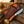 A fixed-blade knife with a patterned brown leather sheath and wooden handle, displayed on rustic wood with a rope and a blurred outdoor western background.
