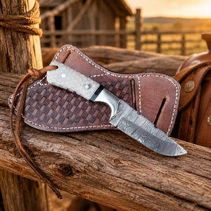 A fixed-blade knife with a patterned steel blade and textured handle rests on a brown leather sheath with a woven center panel, placed on a rustic wooden post outdoors at sunset.