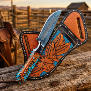 A custom knife with a patterned steel blade and a hand-tooled brown handle, displayed in a matching brown leather sheath with blue and brown floral inlay, set on a rustic wooden log with a blurred western background.
