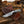 A fixed-blade knife with a patterned steel blade and textured handle, resting on a brown leather sheath over a rustic wooden surface, with a cowboy hat and outdoor fence blurred in the background.
