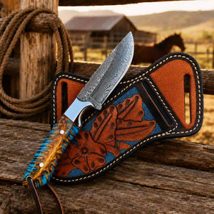 A fixed-blade knife with a patterned steel blade, a blue and tan wrapped handle, and a brown leather sheath with floral tooling, displayed on a rustic wooden surface with a rope and barn in the background.