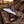 A fixed-blade knife with a patterned steel blade and a carved bone handle rests on a brown leather sheath, placed on a rustic wooden surface with a rope and a leather saddle in a blurred outdoor setting.