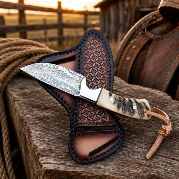 A fixed-blade knife with a patterned steel blade and a carved bone handle rests on a brown leather sheath, placed on a rustic wooden surface with a rope and a leather saddle in a blurred outdoor setting.