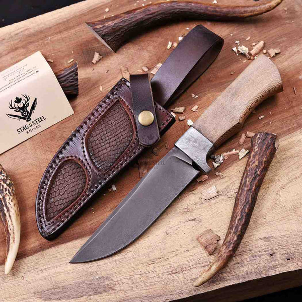 A fixed-blade knife with a light wood handle and dark metal blade, shown beside a brown leather sheath on a wooden surface with antlers and wood shavings. A card reading STAINLESS STEEL Knives is visible in the upper left.