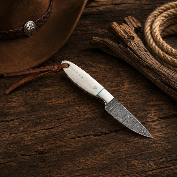 A small fixed-blade knife with a patterned steel blade and a white handle with a blue accent line, resting on a rustic dark wood surface. A brown cowboy hat, leather strap, wood pieces, and rope are styled around it.