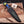 A custom handcrafted knife with a marbled blue, green, and gold handle, resting on a black leather sheath on a rustic wooden board with wood shavings and antlers.