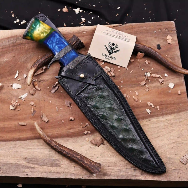 A custom handcrafted knife with a marbled blue, green, and gold handle, resting on a black leather sheath on a rustic wooden board with wood shavings and antlers.