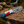 A custom outdoor knife with a patterned Damascus steel blade and a colorful handle, resting on a weathered log with a forest and waterfall background.
