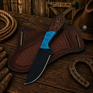 A fixed-blade knife with a textured black blade, a bright blue handle insert, and a dark wood handle, displayed on a brown leather sheath over a rustic wooden surface with western-themed props including a rope, hat, gloves, and horseshoe.