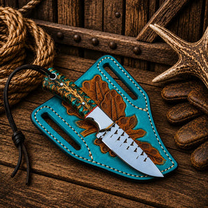 A fixed-blade knife with a silver blade and a carved wooden handle rests on a turquoise leather sheath with brown leaf embroidery, set on a rustic wooden surface with rope, stones, and antler props.