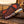 A fixed-blade knife with a patterned tan and blue handle rests on a rustic wooden surface next to a matching brown leather sheath with black stitching, set against a blurred outdoor background at sunset.