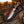 A fixed-blade knife with a textured silver blade and a carved handle lies inside a decorative brown leather sheath on a rustic wooden surface. The background shows a blurred outdoor ranch scene at sunset with a horse and fence.